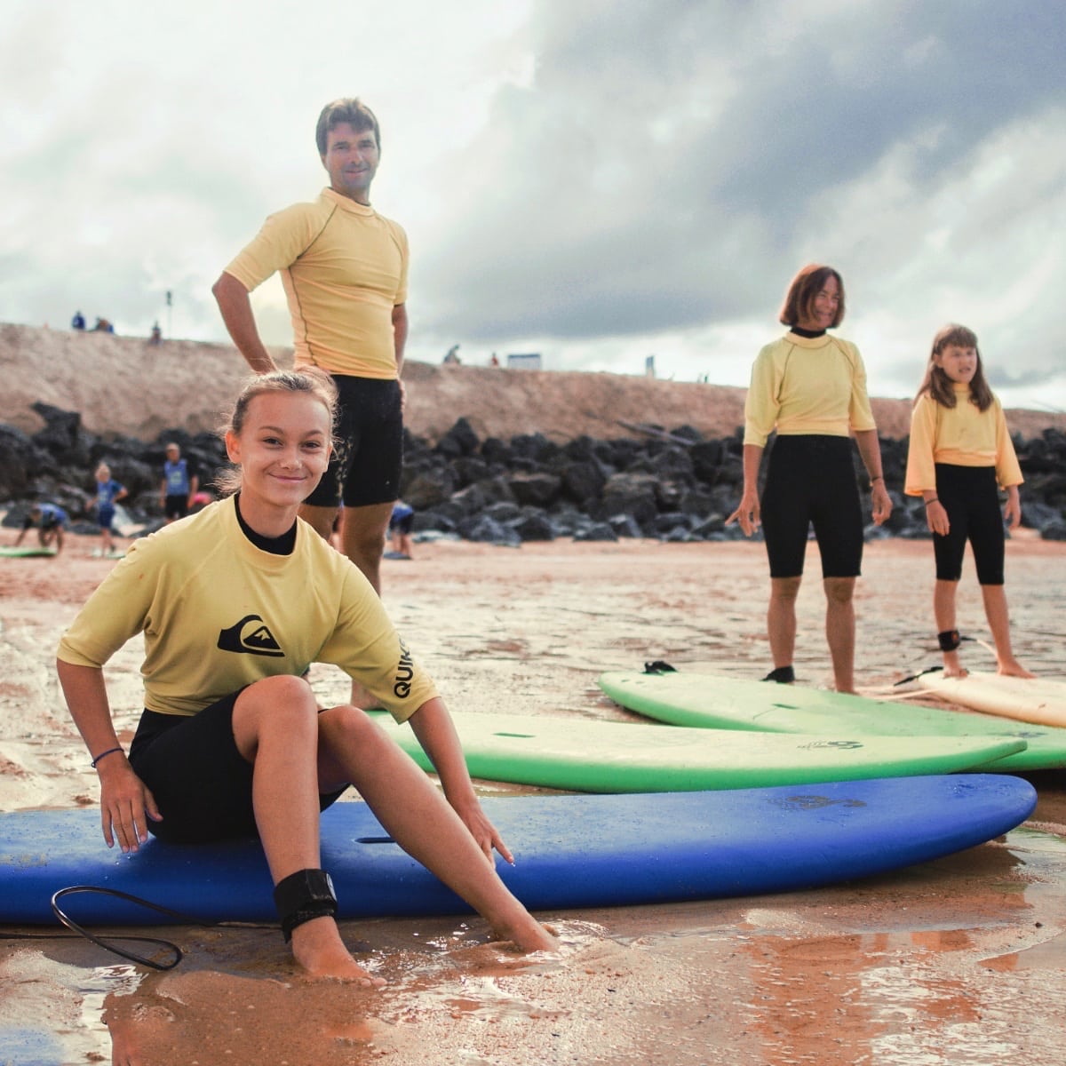 Un groupe de 4 personnes se tient sur la plage avec leurs planches de surf et sourient en regardant la caméra.