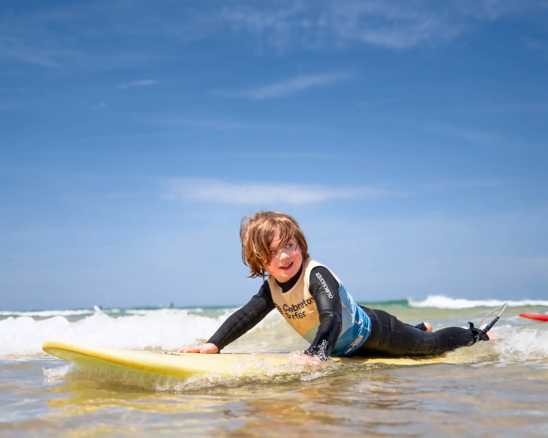 Un enfant allongé sur une planche de surf jaune, souriant, en train d'apprendre à surfer dans des eaux peu profondes, illustrant une scène typique des cours de surf pour enfants